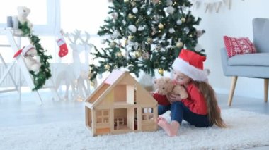 Noel zamanı oyuncak bebek evi ve oyuncak ayıyla oynayan küçük kız Noel Baba şapkası takıyor. Süslü yeni yıl evinde güzel bir çocuk oyuncağıyla