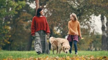 Anne ve kızı sonbahar parkında altın renkli bir köpekle yürüyor. Aile dışarıda köpeklerle dinleniyor..