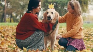 Anne ve kızı sonbahar parkında altın renkli köpekle eğleniyorlar. Mutlu aile dışarıda köpeklerle oynuyor..