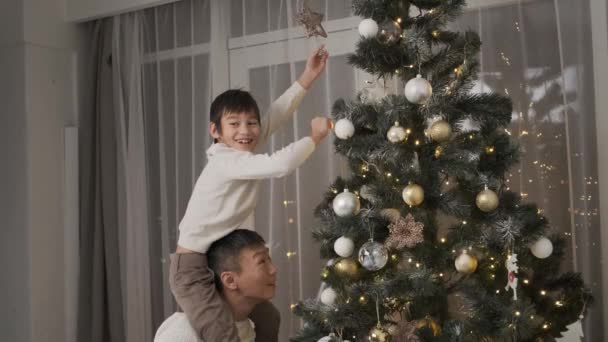 幸せな遠く持ち上げる小さな息子にトップスター上のクリスマスツリーに自宅 — ストック動画
