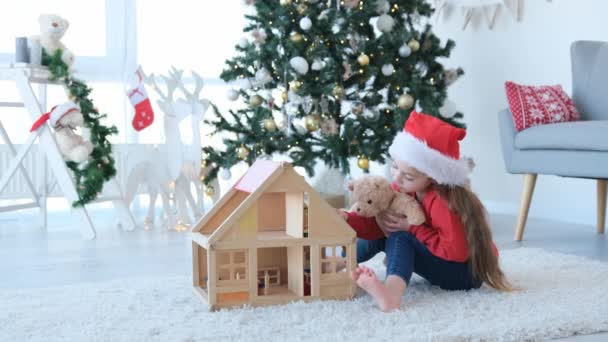 圣诞节的时候 小女孩戴着圣诞老人的帽子 玩木娃娃屋和玩具熊 漂亮的孩子在装饰新年的家与玩具 — 图库视频影像