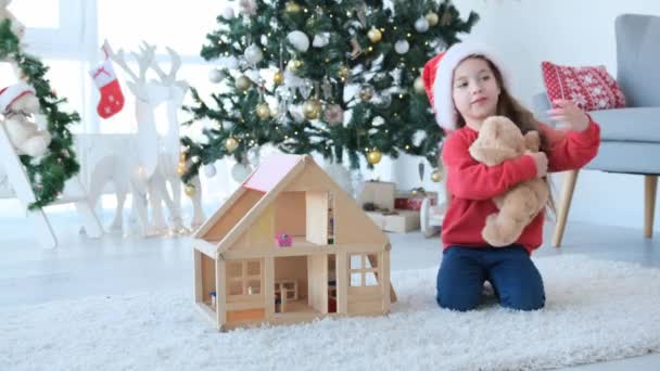 小さな子供の女の子はサンタの帽子をかぶってクリスマスの時間に木製の人形の家の近くにテディベアを抱いています かわいいです子供で装飾された新年の家おもちゃ — ストック動画