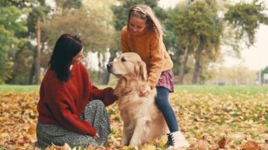Anne ve kızı sonbahar parkında altın renkli köpekle eğleniyorlar. Mutlu aile dışarıda köpeklerle oynuyor..