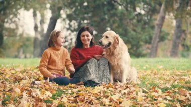 Anne ve kızı sonbahar parkında eğleniyor ve altın renkli köpekleri okşuyor. Mutlu aile dışarıda köpeklerle oynuyor..