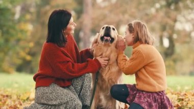 Anne ve kızı sonbahar parkında altın renkli köpekle eğleniyorlar. Mutlu aile dışarıda köpeklerle oynuyor..