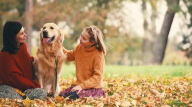 Anne ve kızı sonbahar parkında altın renkli köpekle eğleniyorlar. Mutlu aile dışarıda köpeklerle oynuyor..