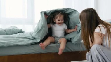 Genç anne pijama giyip yatak odasında küçük kızıyla oynuyor ve gülümsüyor. Genç bir anne ve küçük tatlı bir kız çocuğu gün ışığında odada. Battaniyenin altındaki çocuk