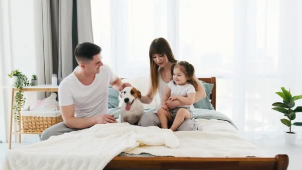 年轻的父母带着小女儿躺在床上 笑着抱着小狗 有小女孩和小狗在房间里的快乐家庭 母亲和孩子早上在一起 — 图库视频影像