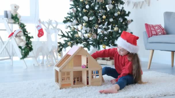 圣诞节的时候 小女孩戴着圣诞老人的帽子在玩木制洋娃娃屋 漂亮的孩子在装饰新年的家与玩具 — 图库视频影像