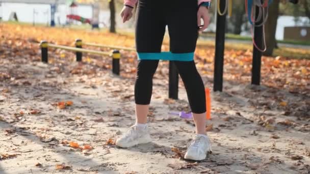 健身女孩在秋季公园里用弹性橡皮筋做蹲 年轻的女运动员在室外进行臀部肌肉运动 — 图库视频影像