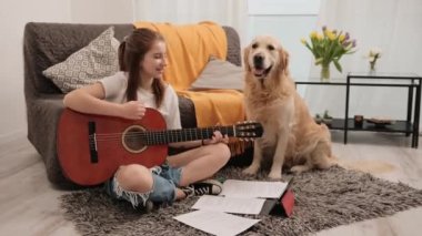 Güzel genç kız evde gitar çalışıyor ve Golden Retriever 'ın köpeğine tutkuyla şarkı söylüyor. Saf köpekle oturma odasında müzik aleti çalma dersi