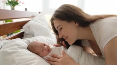 Newborn baby swaddled in white blanket lying in the bed and his young beautiful mother looking at him and smiling. Infant child yawning and his mom cares about him