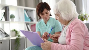 Senior woman and nurse signing documents with pen at home. Young girl medical worker and elderly person preparing medical papers