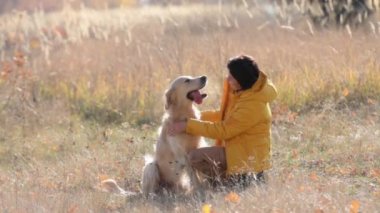 Girl petting golden retriever dog sitting in autumn field and smiling. Purebred doggy with owner at nature outdoors together
