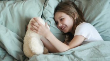 Cute girl looking at teddy bear and smiling in bed under blanket. Little child kid resting with toy in bedroom