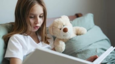 Pretty little girl reading book to teddy bear in bed. Preteen female kid with toy and album at home