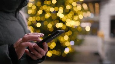 Girl using a smartphone on the street against the background of a Christmas tree with garlands on the street in the city center. On Christmas Eve. New Years City.