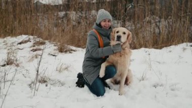 Girl with golden retriever dog sitting in snow park in winter time, petting doggie and smiling. Young woman owner with purebred pet labrador outdoors with cold weather