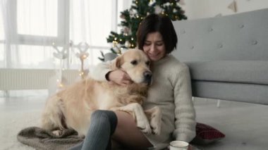Girl petting golden retriever dog and holding cup with cocoa in Christmas time at home with New Year decoration and Xmas lights