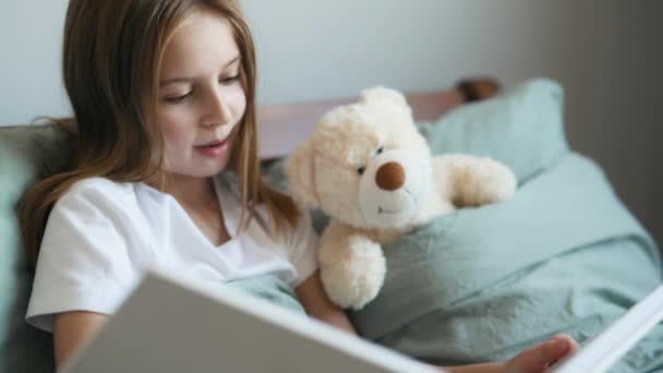 かわいい女の子はベッドの中でテディベアに本のおとぎ話を読んで Preteen女性子供とともにおもちゃとアルバム ホーム — ストック動画