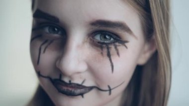 Preteen girl with spooky Halloween makeup and grey green eyes closeup portrait. Creepy child kid with crazy joker expression acting looking at camera