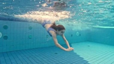 Beautiful girl teenager swimming in pool under water in bikini, side view. Pretty female person diving in blue water at summer