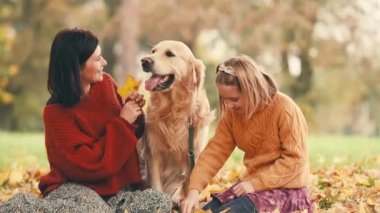 Anne ve kızı sonbahar parkında altın renkli köpekle eğleniyorlar. Mutlu aile dışarıda köpeklerle oynuyor..