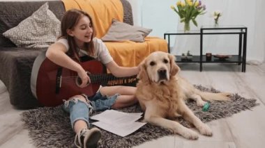 Evde gitar tutan ve Golden Retriever köpeği besleyen güzel bir genç kız. Dişi insan ve safkan köpek müzik aleti çalma dersi sırasında oturma odasında.