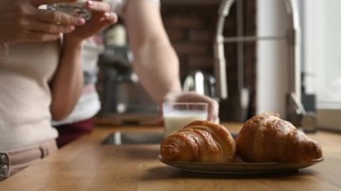 Mutfakta kahvaltıda süt bardaklı ve kruvasanlı kız ve adam. Beyaz organik latte ve Fransız pastası olan kadın ve erkek çift.