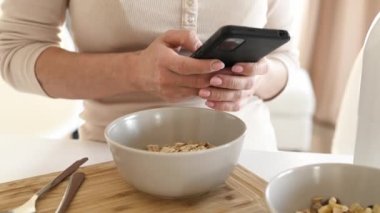 Girl with oatmeal bowl and smartphone in hands checking social media news during cereal breakfast. Woman with mobile phone and granola at kitchen