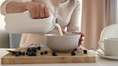 Kız yulaf ezmesine yulaf ezmesi ve yaban mersinli tabağa süt koyar. Mutfakta lezzetli kahvaltılık gevrek için. Sabah sağlıklı diyet yemeği için meyveli ve latte hazırlayan kadın.