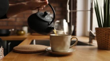 Mutfakta çaydanlıktan sıcak çay dolduran bir kız. Kadın seramik bardağı çaydanlıktan çıkan sıcak bitkisel içecekle dolduruyor.