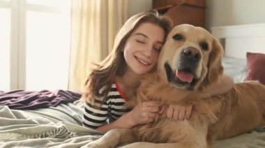 Golden Retriever köpeğini kucaklayan güzel kız yatakta uzanıyor ve gülümsüyor. Safkan evcil köpek labrador 'u olan güzel bir genç kız evde dinleniyor.
