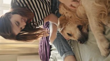 Yatakta Golden Retriever köpeğiyle oynayan ve gülümseyen güzel bir kızın dikey videosu. Safkan evcil köpek labrador 'u evinde dinleniyor.