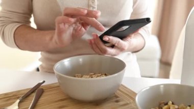 Girl with oatmeal bowl and smartphone in hands checking social media news during cereal breakfast. Woman with mobile phone and granola at kitchen