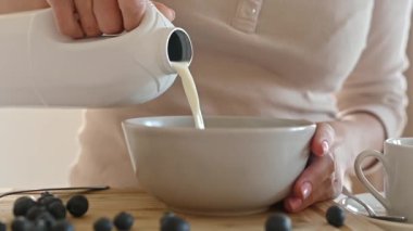 Girl puts milk to oatmeal with blueberry for delicious cereal breakfast at home. Woman preparing granola with berry and latte for morning healthy diet meal