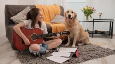 Güzel genç kız evde gitar çalışıyor ve Golden Retriever 'ın köpeğine tutkuyla şarkı söylüyor. Saf köpekle oturma odasında müzik aleti çalma dersi