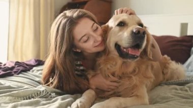 Golden Retriever köpeğini kucaklayan güzel kız yatakta uzanıyor ve gülümsüyor. Safkan evcil köpek labrador 'u olan güzel bir genç kız evde dinleniyor.