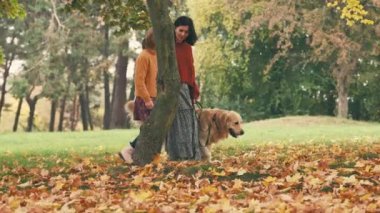 Anne ve kızı altın renkli bir köpekle sonbahar parkında yürüyüp konuşuyorlar. Aile dışarıda köpeklerle dinleniyor..