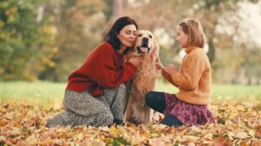 Anne ve kızı sonbahar parkında altın renkli köpekle eğleniyorlar. Mutlu aile dışarıda köpeklerle oynuyor..