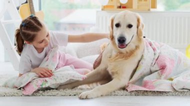 Gülümseyen küçük kız ve sakin köpek oyun odasında battaniyenin altında yatıyor.