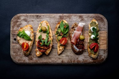 Set of bruschettas with jamon, olives, pesto, grilled and cherry tomatoes, basil served on wooden board with arugula. Traditional mediterranean toasted bread with cheese, meat and vegetables clipart