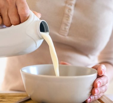 Girl puts milk to oatmeal with blueberry for delicious cereal breakfast at home. Woman preparing granola with berry and latte for morning healthy diet meal