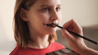 Anne, reşit olmayan kıza Cadılar Bayramı makyajı yapıyor ve yüzüne ürkütücü siyah çizgiler çiziyor. Ürkütücü kozmetik ürünleri olan güzel bir çocuk Ekim tatili partisine hazırlanıyor.