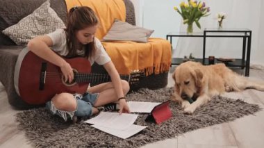 Güzel genç kız evde gitar çalışıyor ve Golden Retriever köpeği yerde yatıyor. Saf köpekle oturma odasında müzik aleti çalma dersi