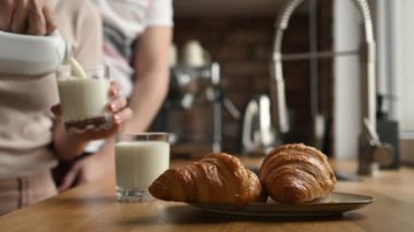 Bardağa süt döken ve mutfakta kruvasanla kahvaltı yapan bir kız. Beyaz organik latte ve Fransız pastası olan kadın ve erkek çift.