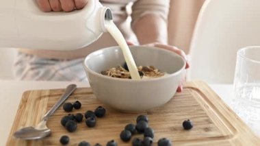 Kız yulaf ezmesine yulaf ezmesi ve yaban mersinli tabağa süt koyar. Mutfakta lezzetli kahvaltılık gevrek için. Sabah sağlıklı diyet yemeği için meyveli ve latte hazırlayan kadın.