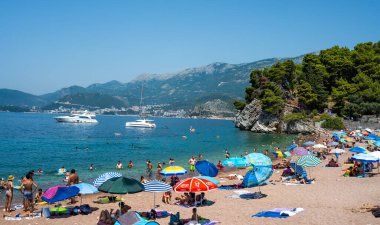 Budva, Karadağ - 27 Temmuz 2021: Karadağ 'ın Budva kentindeki Sveti Stefan adasındaki plaj, güneşli bir günde dağlar manzaralı. Adriyatik denizinde lüks tatil beldesindeki insanlar