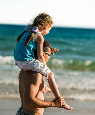 Baba deniz kıyısında kızını omuzlarına alıp uzaklara bakıyor. Aile babası ve küçük tatlı çocuk yazın plajda birlikteler.