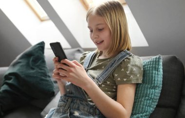 Akıllı telefonlu kız evdeki kanepede oturuyor ve sosyal medyada arkadaşıyla sohbet ediyor. Cep telefonuyla mesajlaşan güzel bir çocuk.